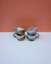 Two ceramic mugs with a striped pattern on a white surface against an orange background. Liz Vidal Ceramics | Shop Fodder