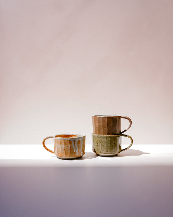 Three handmade ceramic mugs with rustic glazes in earthy tones of brown, green, and orange, arranged on a white surface against a soft beige background. Carved Mug | Scree Ceramics | Fodder