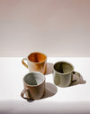 Three handmade ceramic mugs with different glazes in warm earthy tones, arranged on a white surface against a beige background. Everyday Mug | Scree Ceramics | Fodder