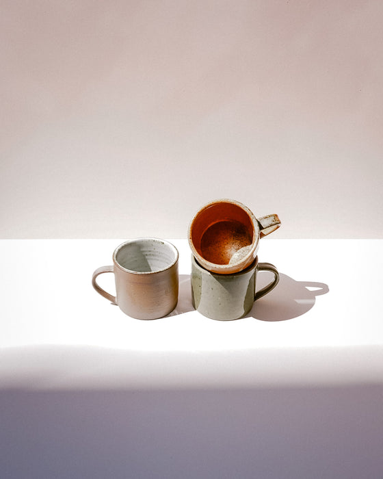 Three handmade ceramic mugs with organic shapes and handles, displayed on a white surface against a warm beige background. One mug is stacked on top of the other, showing a warm terracotta-brown interior, while the others display speckled beige and sage green exteriors. Everyday Mug | Scree Ceramics | Fodder