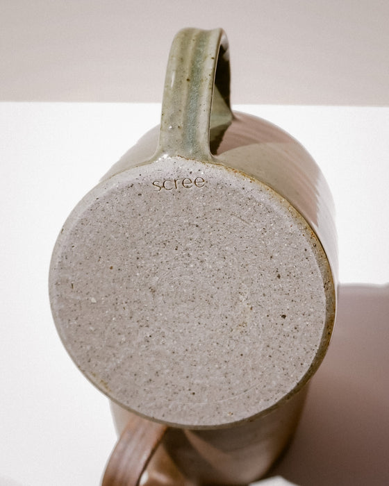 Close-up of a ceramic mug showing the textured, speckled light gray bottom and the handle, with the brand name 'scree' printed on the side, set against a neutral beige background. Everyday Mug | Scree Ceramics | Fodder