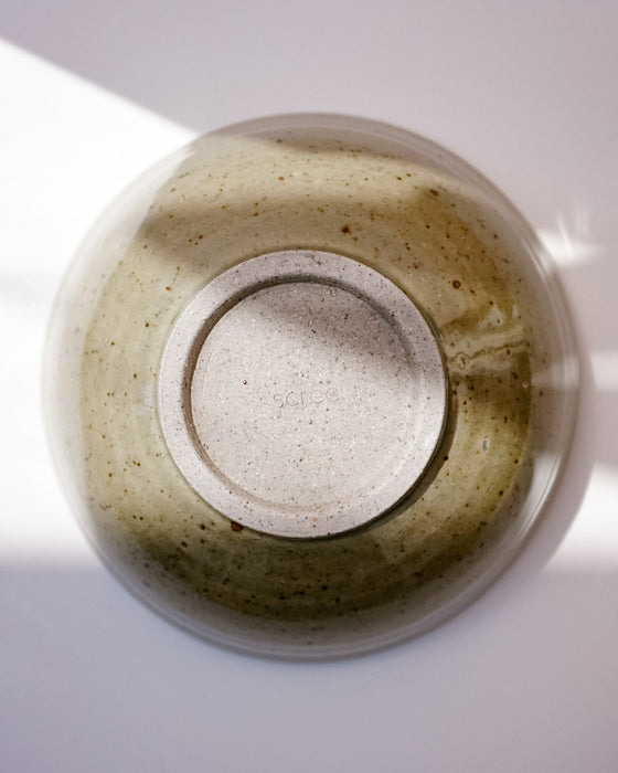 An overhead close-up of a round ceramic bowl with a speckled sage green and beige glaze, photographed on a white surface with soft natural shadows. Morning Bowl | Scree Ceramics | Fodder