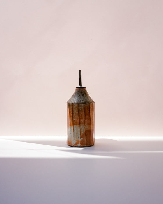 A small ceramic bottle with a conical shape, featuring a textured glaze in orange, brown, and cream, with a dark spout, set against a soft pink background with a white surface below. Oil Bottle | Scree Ceramics | Fodder