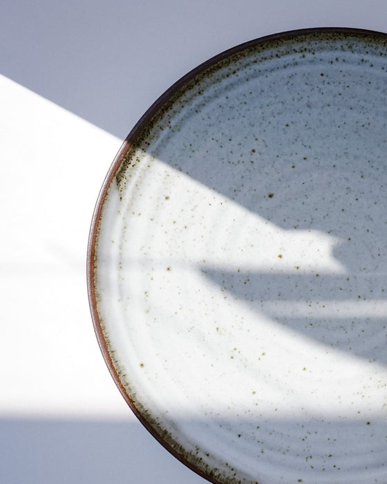 A close-up of a round ceramic plate with a speckled off-white glaze and a dark brown rim, photographed against a light background with strong diagonal shadows. | Scree Ceramics | Fodder