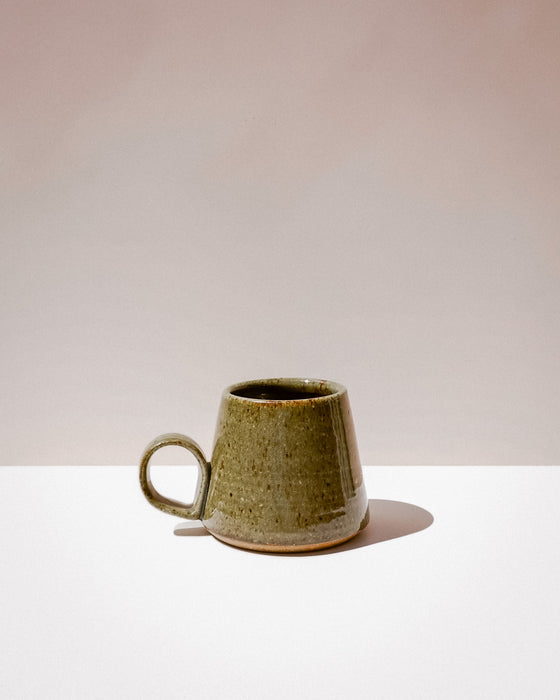 A small handmade ceramic mug with a tapered shape and a loop handle, featuring a textured olive green glaze with brown speckles, set against a minimalist beige and white background. Tall Loop Mug | Scree Ceramics | Fodder