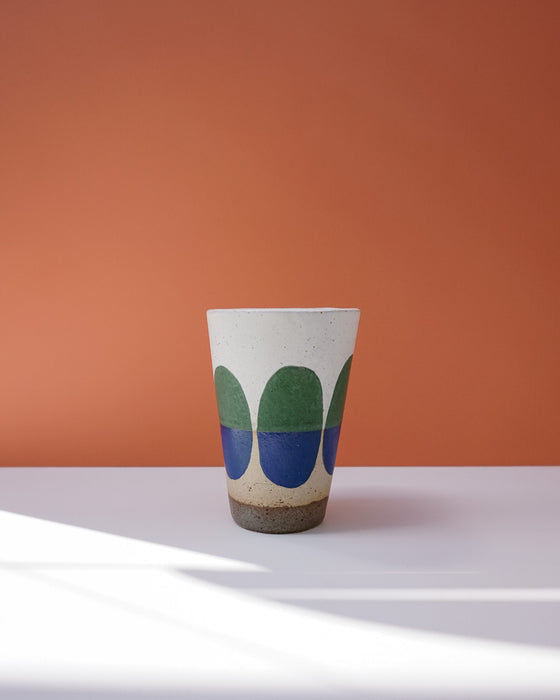 Ceramic cup with green and blue design on a white surface against an orange background. Sophie Alda | Handmade Ceramics | Shop Fodder