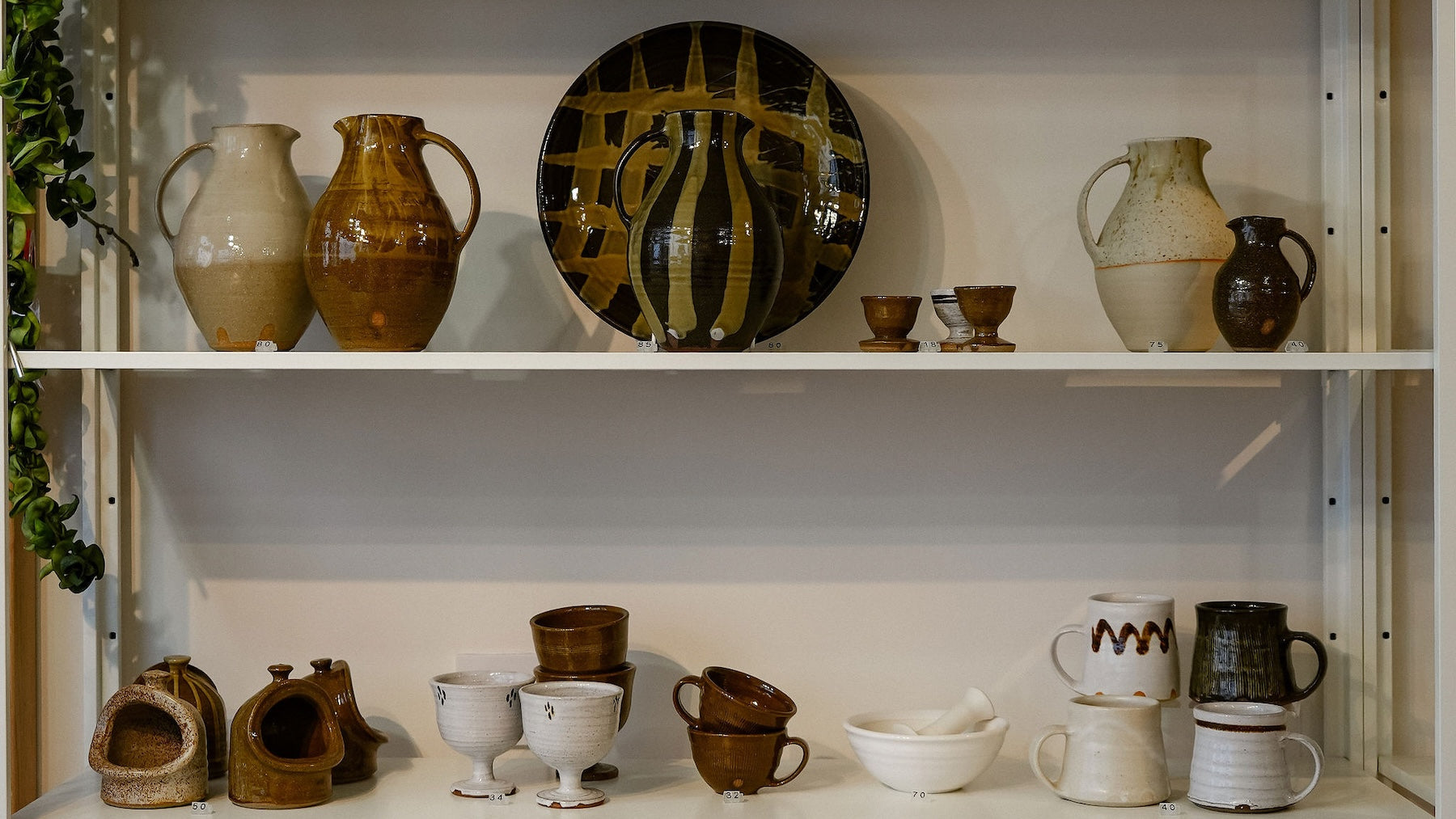 A collection of handmade ceramic pottery, including pitchers, mugs, cups, and a decorative plate, displayed on white shelves.